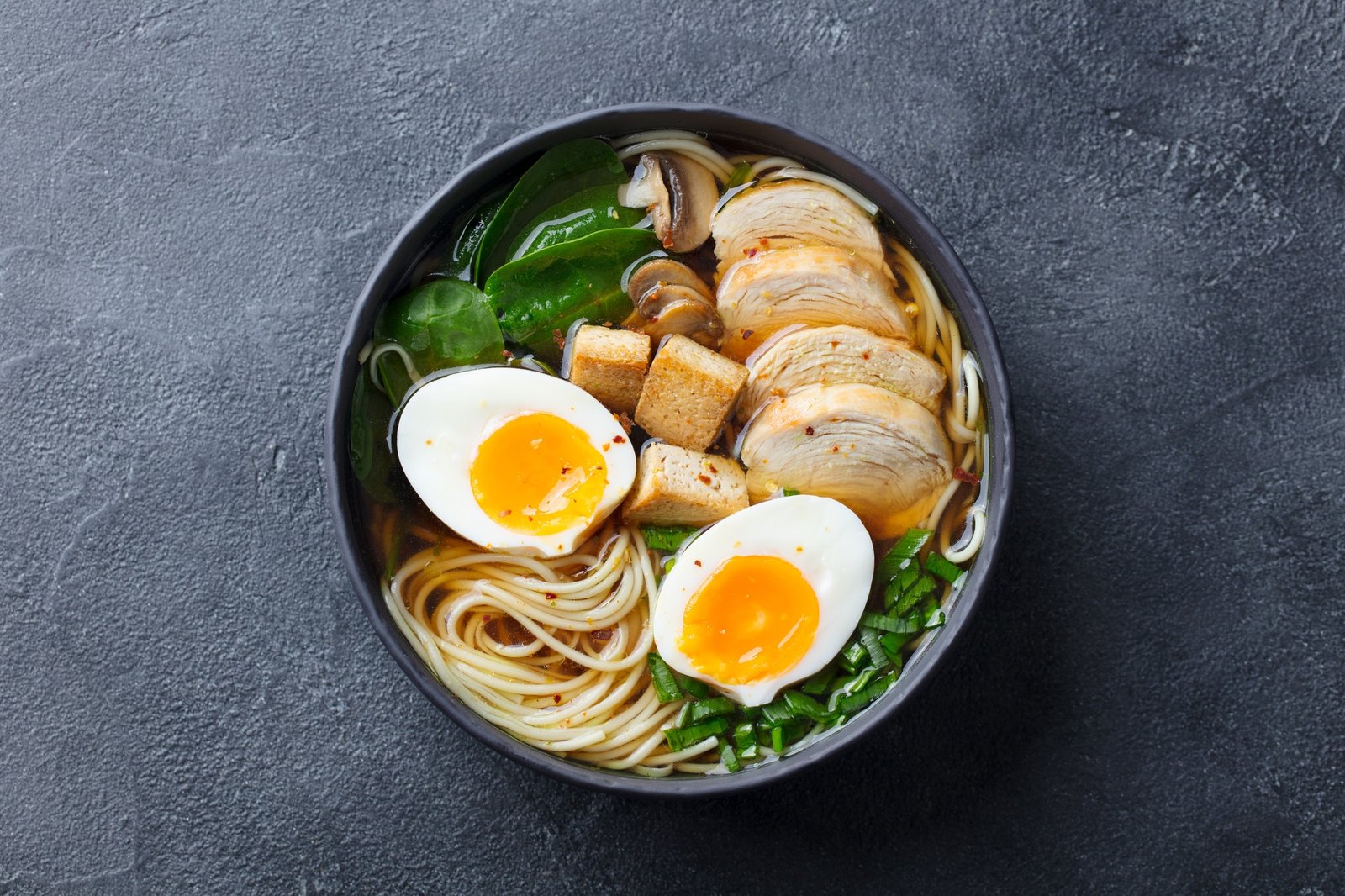 Asian Noodle Soup, Ramen with Chicken, Tofu, Vegetables and Egg in Black Bowl. Slate Background.